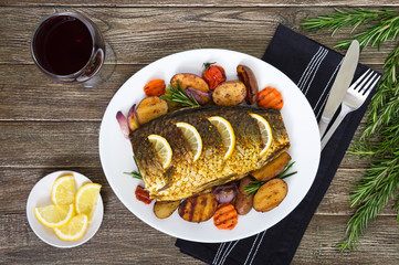 Baked carp with vegetables grilled on a white plate and a glass of red wine on a wooden background. Top view.