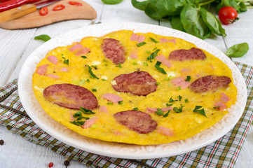 Omelette with ham, salami, cheese and greens on a plate on a white wooden background. Breakfast.