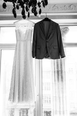 Bridal dress and a groom’s suit on the hangers on a window. Wedding concept. Black and white photo.