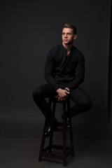 A white man sits on a dark chair in a black shirt and black mills, on a gray background