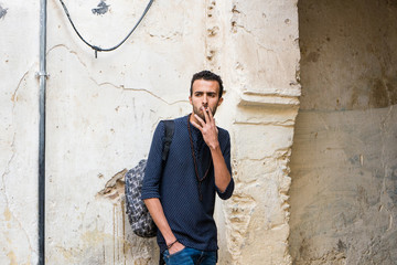 Young Muslim man smoking a cigarette in casual clothing standing by the ruined wall