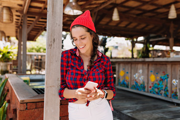 Beautiful lovely young woman with dark hair dressed in plaid shirt and red cap poses with shy...
