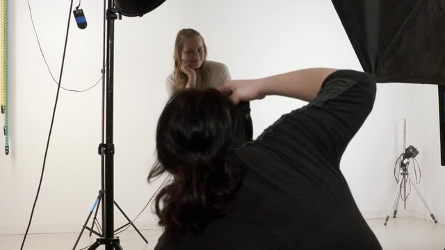 Studio photography. Female photographer working in photo studio with model. Young blonde girl poses for photographer on white background. Backstage shot during indoor photoshoot.