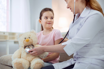 Obraz na płótnie Canvas Great patient. Pretty little girl looking at female doctor who sitting next to her and treating plush bear