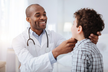 Pain in throat. Handsome afro American male doctor looking at boy while smiling and checking his...