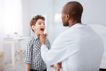 Obraz premium Strep throat. Pretty nice boy standing on blurred background while showing throat to doctor and waiting for instructions