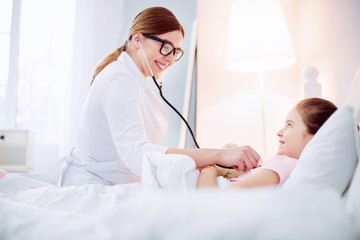 Obraz premium Good doctor. Optimistic vigorous female doctor hearing girl with stethoscope while smiling and looking at her
