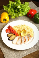 Grilled chicken with slices, with vegetables, bulgur on a white plate. In the background, lettuce leaves, color pepper. Low-calorie, dietary food for a healthy lifestyle. Wooden table, burlap