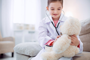 Plush friend. Little kind peaceful girl listening to plush bear while using stethoscope and sitting on couch