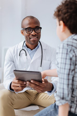 Digital data. Gay afro American male doctor wearing glasses while showing tablet to boy and sitting