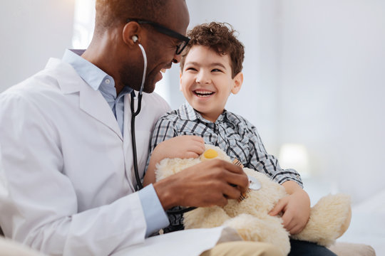 Immediate treatment. Enthusiastic vigorous male doctor sitting with boy while listening to plush bear and talking