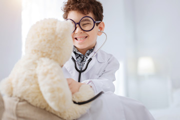 Future doctor. Jovial happy satisfied boy wearing glasses while listening to plush bear and grinning