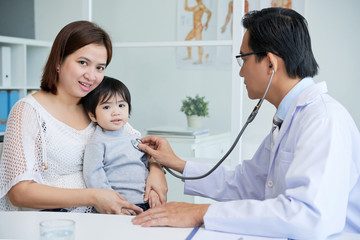 Profile view of highly professional doctor wearing white coat using stethoscope in order to examine little patient, his mother looking at camera with wide smile