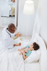 Little fun. Top view of afro American experienced male doctor gesturing while boy lying in bed and hugging bear