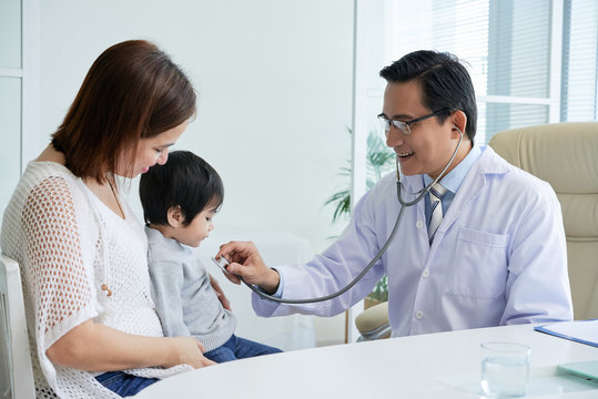 Middle-aged doctor looking at his cute little patient with wide smile while examining him with stethoscope, interior of modern office on background