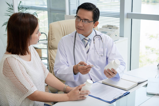 Highly professional Asian physician wearing eyeglasses sitting at desk and explaining pretty young patient how to take pills correctly, interior of modern office on background