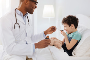 Obraz premium Cough medicine. Joyful merry boy laughing while male doctor looking at him and stretching spoon with medication