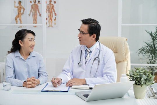 Smiling senior patient sitting at desk next to middle-aged physician and answering questions while he filling in medical record, interior of modern office on background
