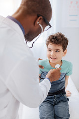 Cunning look. Male doctor standing while using stethoscope and auscultating jovial merry boy