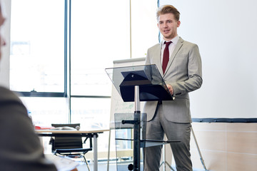 Portrait of smiling young businessman standing by podium while giving speech at conference, copy...