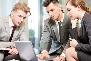 Group of three young business people using laptop and collaborating on project sitting at coffee table in office