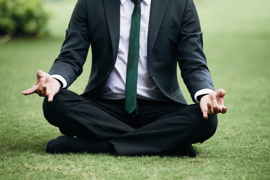 Unrecognizable businessman in sut meditating on green grass in park