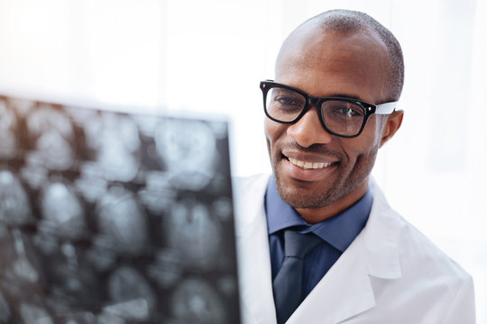Brain issue. Jolly merry male doctor wearing glasses while grinning and looking at brain imaging