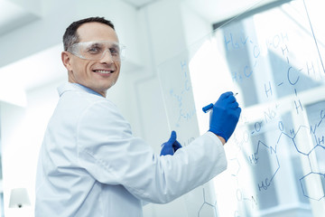 Feeling happy. Handsome alert dark-haired smart scientist smiling and wearing a uniform and writing formulas