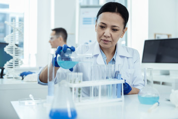 New experiment. Pretty concentrated smart dark-haired scientist holding a glass and making an experiment and her colleague sitting in the background