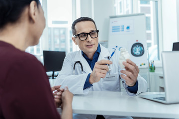 Want to help. Professional pleasant therapeutist sitting at the table and holding a model of heart