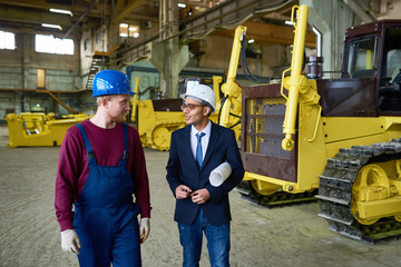 Portrait of young Middle-Eastern engineer talking to factory worker walking across industrial workshop with tractor, copy space