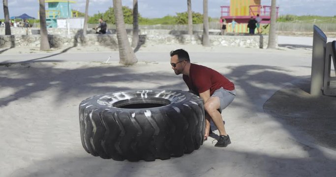 Side view of strong athletic man in sunglasses and sportswear pushing huge heavy tyre in bright sunlight on sandy beach. 