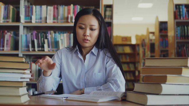Young asian teenage girl is sitting at desk in big library rewriting text from book. She is tired and exhausted, looking through text closing book and turning away