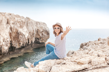 Naklejka na ściany i meble Young beautiful woman travel girl in hat and sunglasses sits on a rocky seashore, summer vacation and travel, sunny portrait