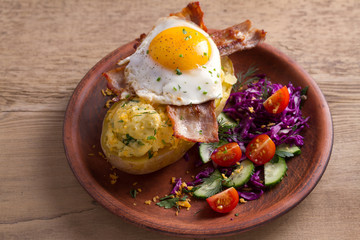 Baked potato in jacket loaded with cheese and topped with bacon and fried egg on plate with vegetables. Stuffed potato with topping