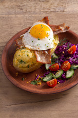 Baked potato in jacket loaded with cheese and topped with bacon and fried egg on plate with vegetables. Stuffed potato with topping