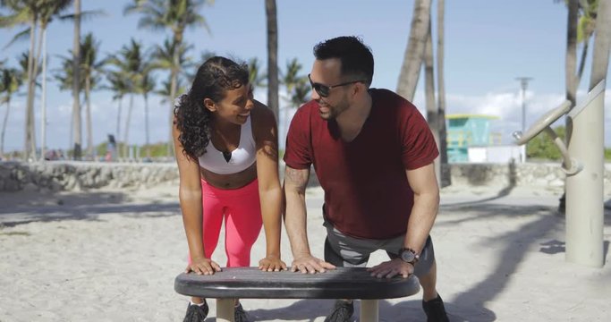 Laughing black woman and man in sportswear working out happily on sandy beach with gym in sunlight. 