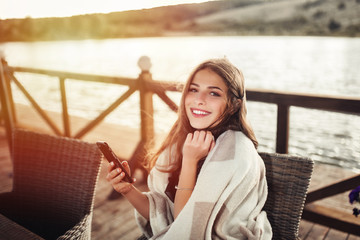 Cheerful relaxed woman uses smart phone for chat with friends, sits in modern coffee shop or terrace cafe. Pretty female reads good news on internet website, enjoys summer rest.