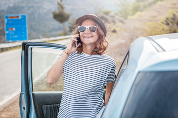 Young beautiful female travel girl in hat and sunglasses speaks by mobile phone on the background of her car while traveling in the mountains, renting and car service