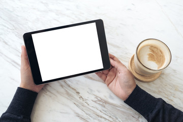 Top view mockup image of hands holding black tablet pc with white blank screen and coffee cup on table background