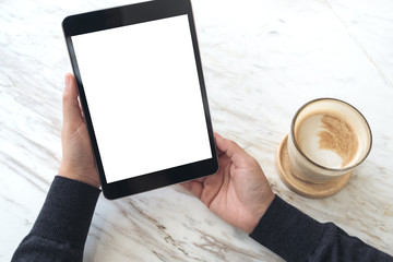Top view mockup image of hands holding black tablet pc with white blank screen and coffee cup on table background