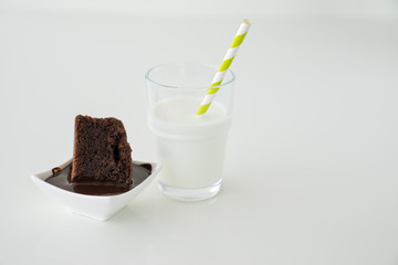 Slice of delicious homemade chocolate brownie served on white plate. Served with glass of milk with green white stripe straw. Taken on a white background.