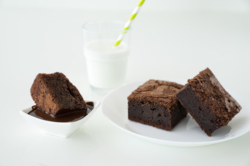 Slice of delicious homemade chocolate brownie served on white plate. Served with glass of milk with green white stripe straw. Taken on a white background.