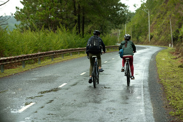 Rear view of couple cycling together down wet road, man wearing backpack