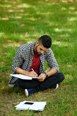 Naklejka na ściany i meble Concentrated Indian student in casualwear sitting in lotus position on green lawn and taking notes while preparing for university exam
