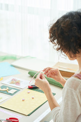 Profile view of curly woman in casualwear sitting at wooden table while enjoying process of handmade greeting card creation