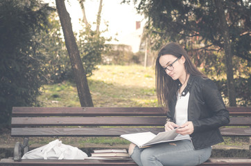 Obraz premium Young beautiful school or college girl with eye glasses sitting on the bench in the park reading the books and study for exam, knowledge and education concept