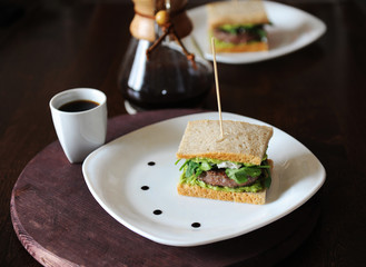 Fototapeta na wymiar Burger with beef and greens in slices of unleavened bread. On white plate with drops of sauce. Coffee on background
