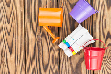 Heap of colorful acrylic paints in closed containers and two empty metal buckets and orange watering can on old wooden brown boards. Top view