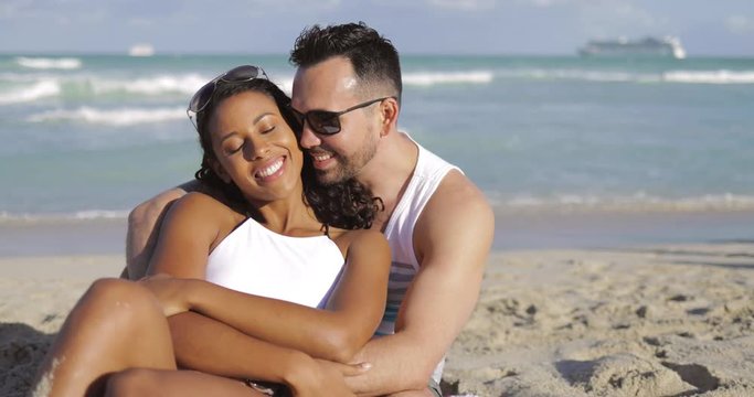 Happy multiracial man and woman embracing while sitting on white sand of beach in sunlight on background of ocean waves. 